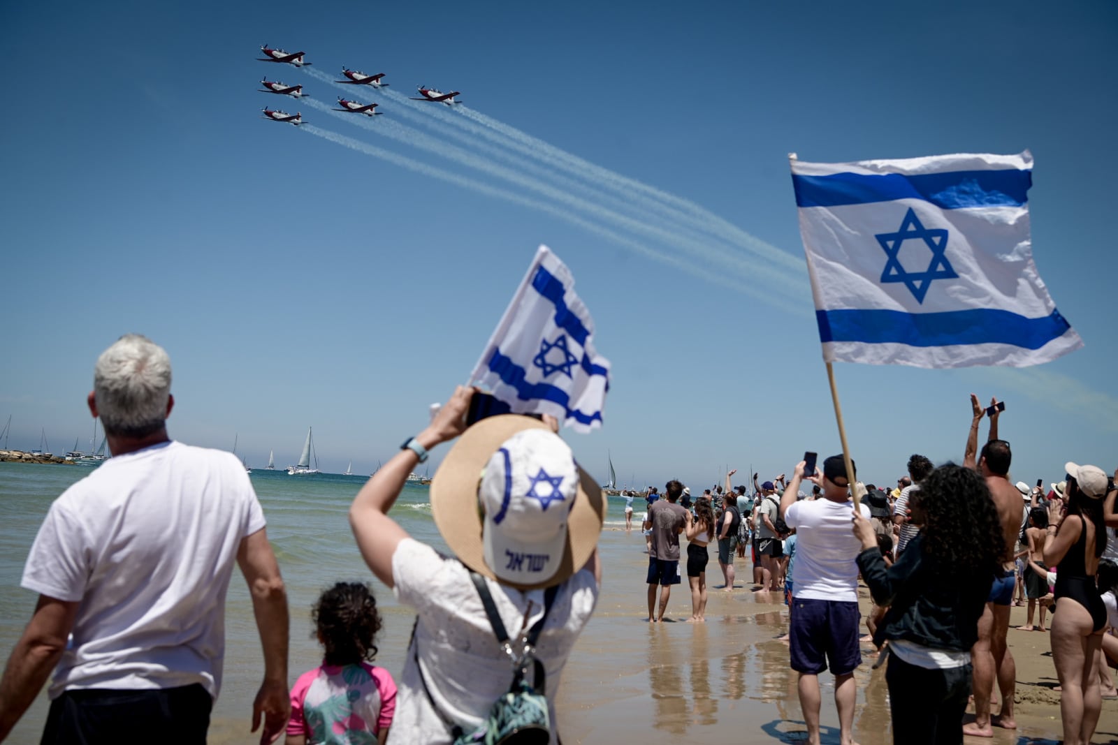 People stand on a beach waving Israeli flags and watching a formation of airplanes flying overhead, leaving trails in the sky on Israel's independence day. Some are taking photos, and boats are visible on the water under a clear blue sky.