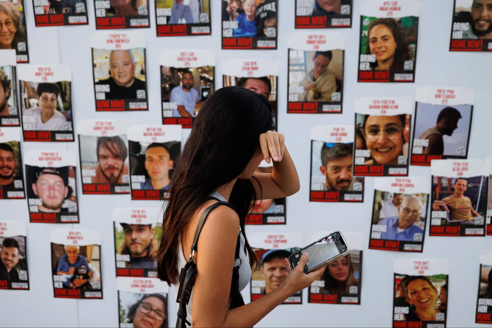A woman holding a smartphone stands in front of a wall displaying photos of hostages taken on October 7. She dries tears in her eyes with her hand, looking closely at the images arranged in a grid pattern on a white background.