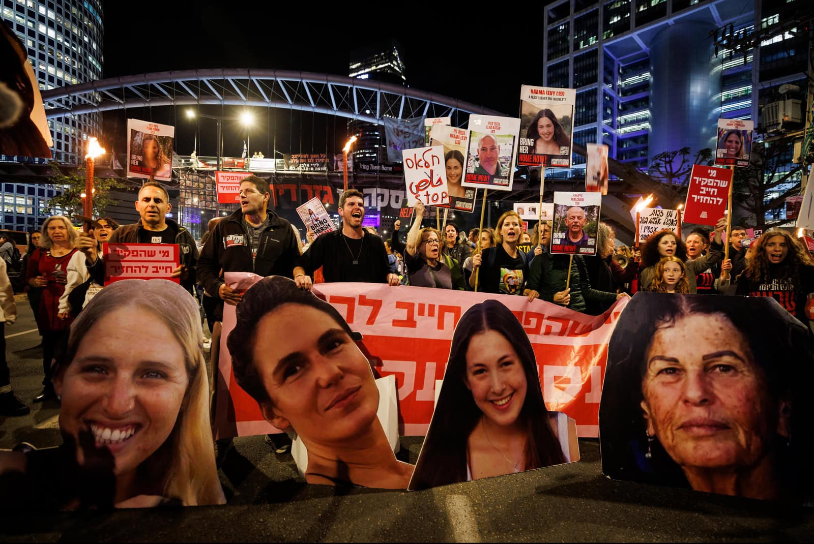 A nighttime demonstration in Tel Aviv where protesters hold large photos of Israeli hostages with text. A variety of expressions are visible in the crowd, and there are bright lights and a bridge in the background. 