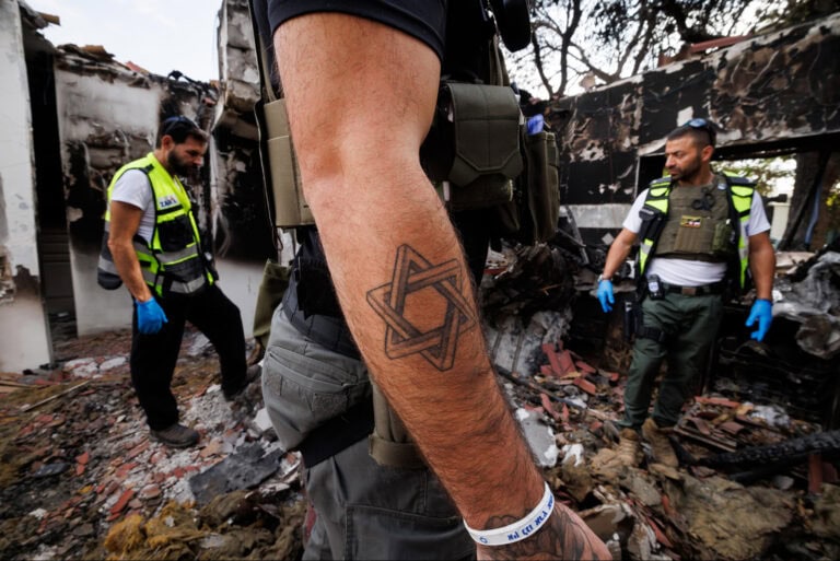 Three ZAKA volunteers in tactical gear inspect a damaged, debris-filled area. One person's arm, adorned with a Star of David tattoo, is prominently visible in the foreground. Burnt structures and rubble are in the background. 