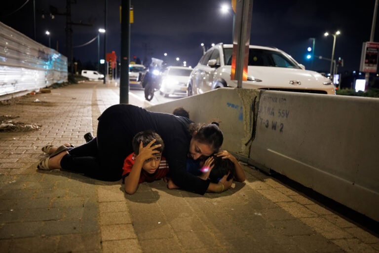 A woman is lying on a sidewalk shielding her two children with her body. Vehicles are stopped on the road nearby, and its nighttime. The scene is tense, with the woman holding the children closely under her arms. 