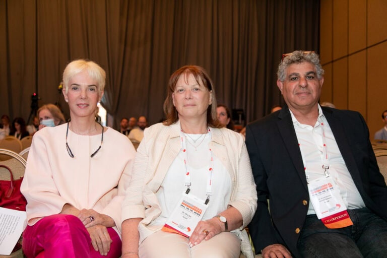BioMed Israel co-chairs, from left, Ruti Alon, Ora Dar and Nissim Darvish, sit in a conference room. They are wearing professional attire and have conference badges around their necks. The background shows a partially visible audience and beige curtains.