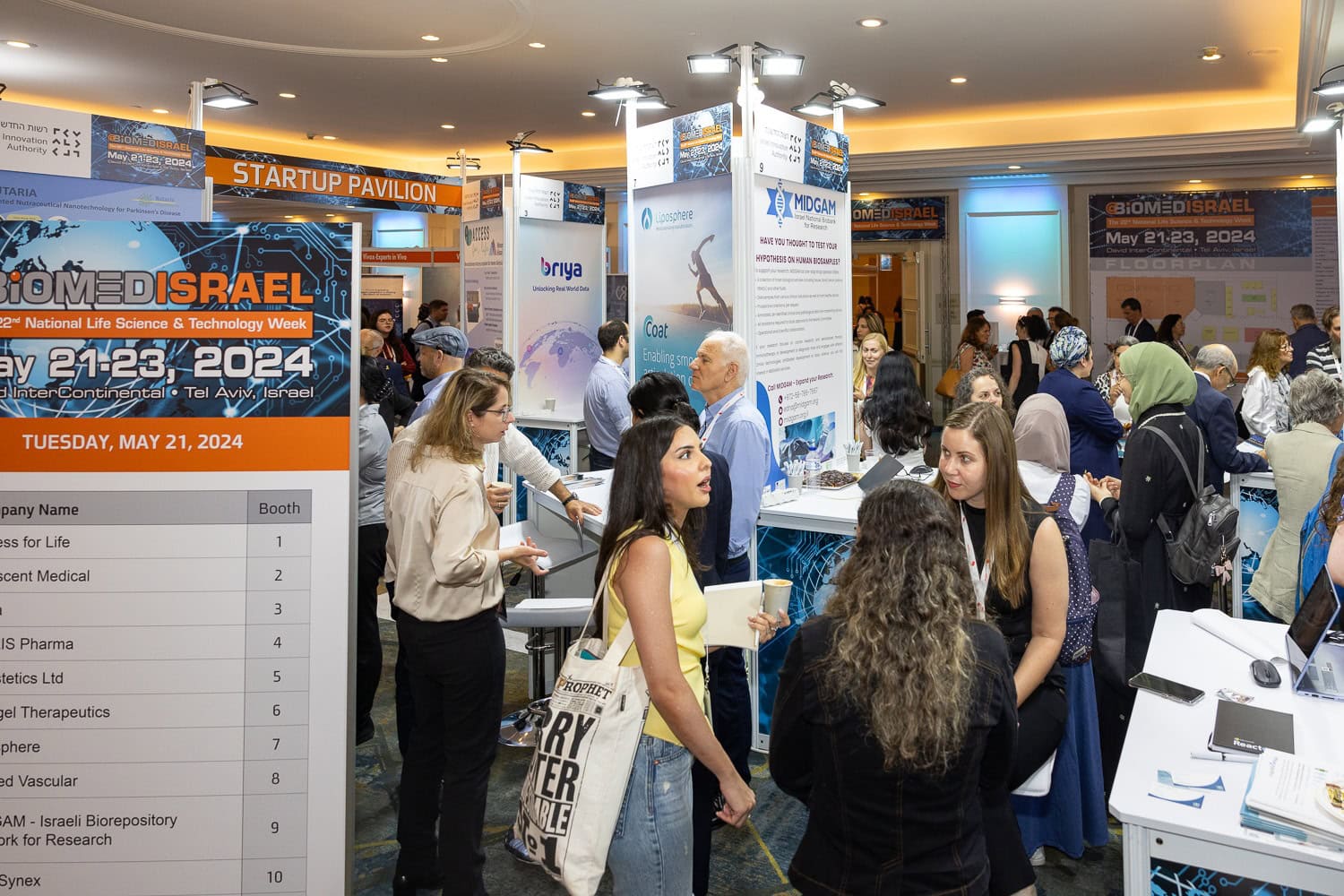 People are gathered in a busy exhibition hall for the BioMed conference. Booths display information and banners about medical science and technology. A large poster mentions the event dates, May 21-23, 2024.