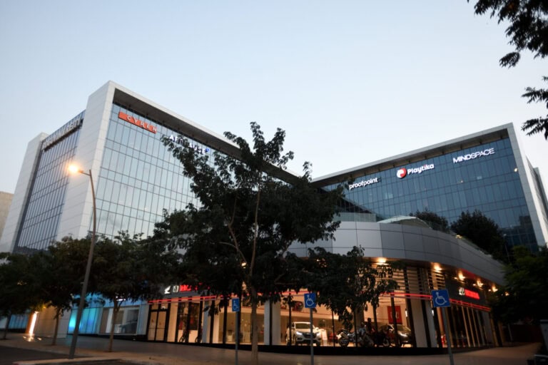 Two large buildings covered in windows, with a parking lot at the bottom of the building and a grey sky in the background. The lights are on and the buildings are surrounded by trees. 