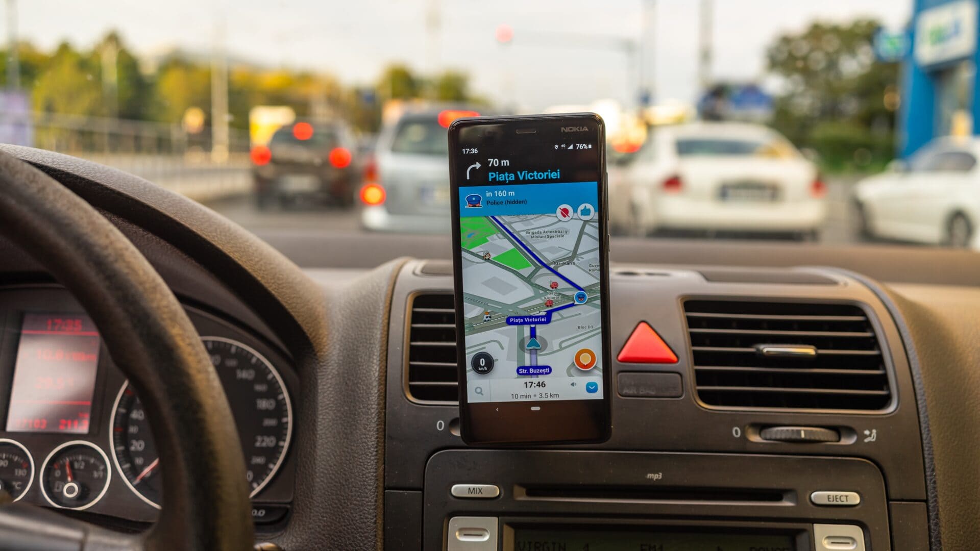 A car dashboard with a phone attached to it. The phone screen shows a digital map and directions. In the background, we see a blurry view of ongoing traffic in front of the care. 