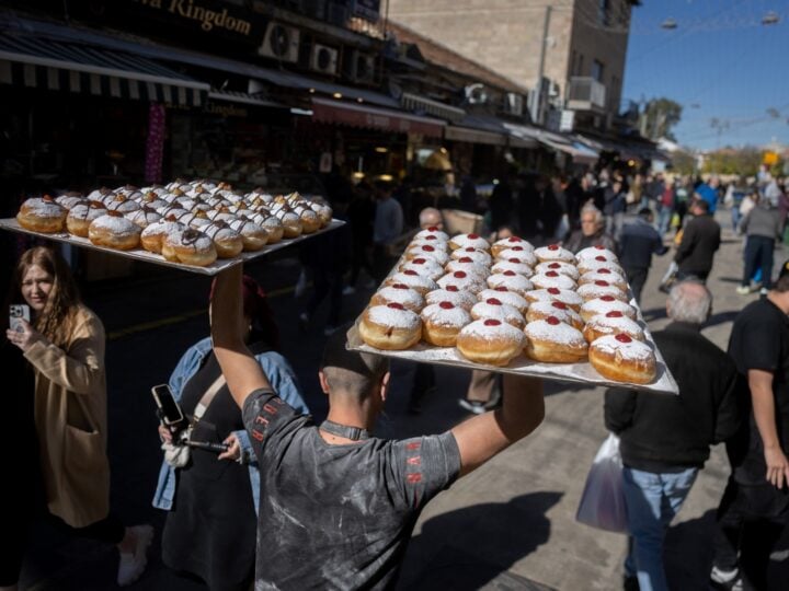 A vendor carries two large trays of jelly-filled, powdered donuts above his head while walking through the crowded Machne Yehuda market in Jerusalem. People walk and shop along the busy street under a clear sky during Hanukkah.
