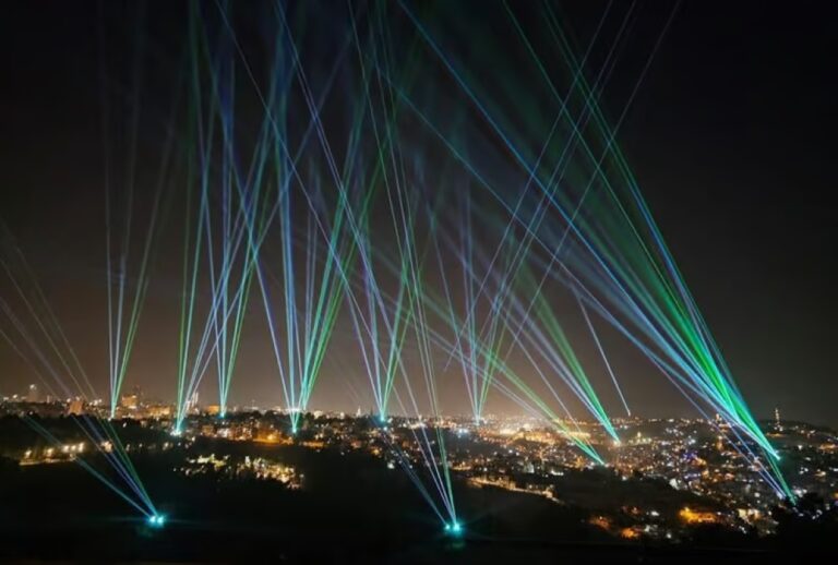 Colorful beams of blue and green laser lights shoot up into the night sky over a cityscape, illuminating buildings and creating a vibrant, festive atmosphere.
