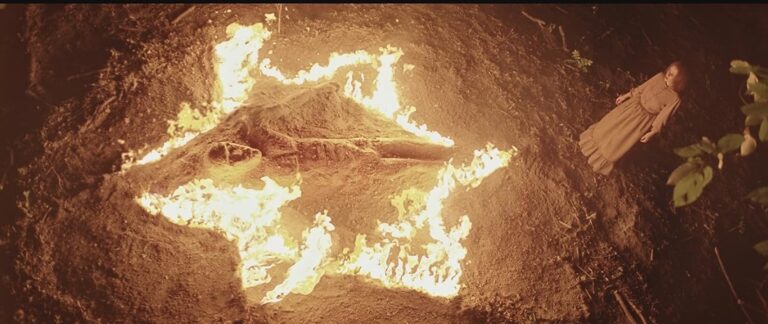 A woman in a long dress stands near a burning fire in the shape of a five-pointed star, with a large object or figure at the center, surrounded by dirt and foliage, viewed from above at night.
