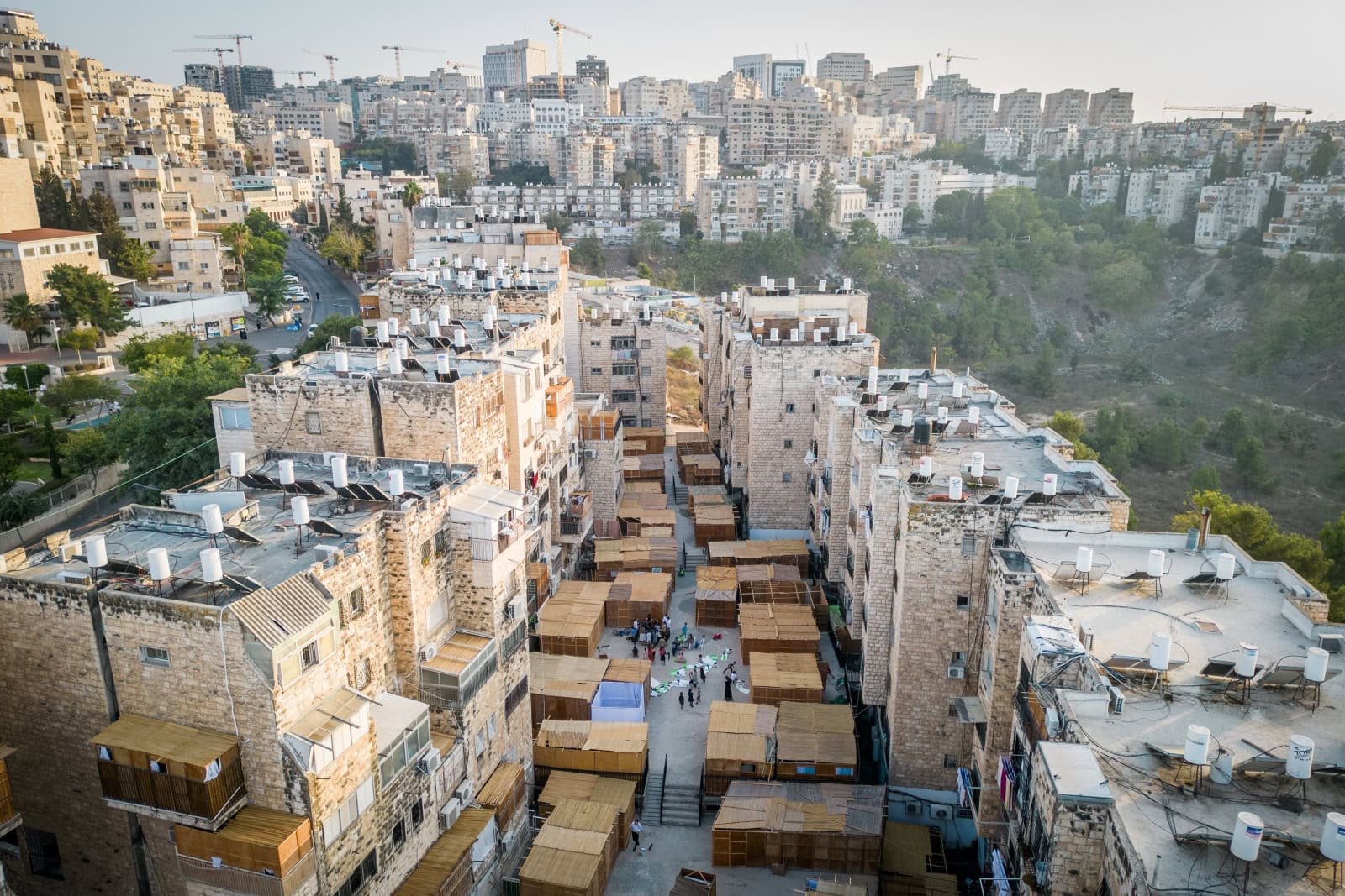Aerial view of a densely built urban area in Jerusalem during the holiday of Sukkot. We see tall apartment buildings and rows of Sukkot, which are temporary wooden structures, set up in the courtyards below. The street and buildings are surrounded by greenery and a hillside in the background.