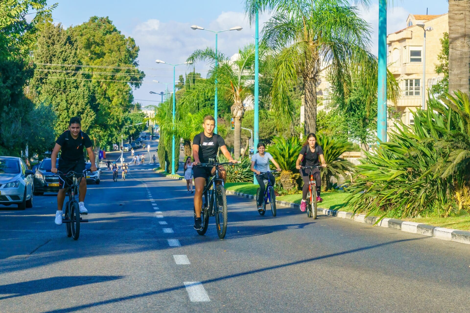 Four Israelis ride bicycles on a sunny, tree-lined city street on Yom Kippur, wearing casual athletic clothing. Two cyclists are on the left and two on the right, with green plants and buildings in the background.