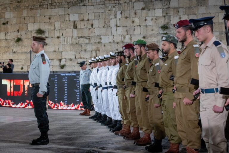 A line of uniformed soldiers stands in formation in front of the Western Wall, with some dressed in white and others in brown and tan uniforms, during a formal ceremony at night.