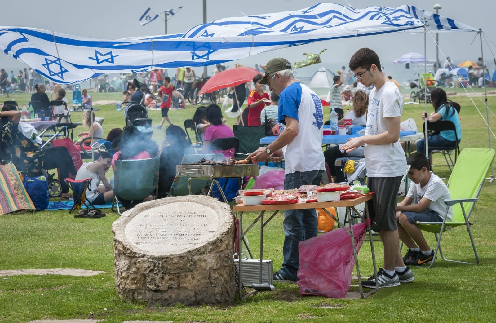 People gather outdoors for a picnic, with two men grilling near a table. Children and families sit on lawn chairs under tents with Israeli flags. The scene is lively and festive, with a tree stump in the foreground.