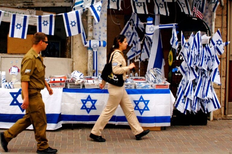 A man and a woman in military uniform walk past a street stall decorated with numerous Israeli flags and blue-and-white items, including banners, hats, and tablecloths with the Star of David printed on it.