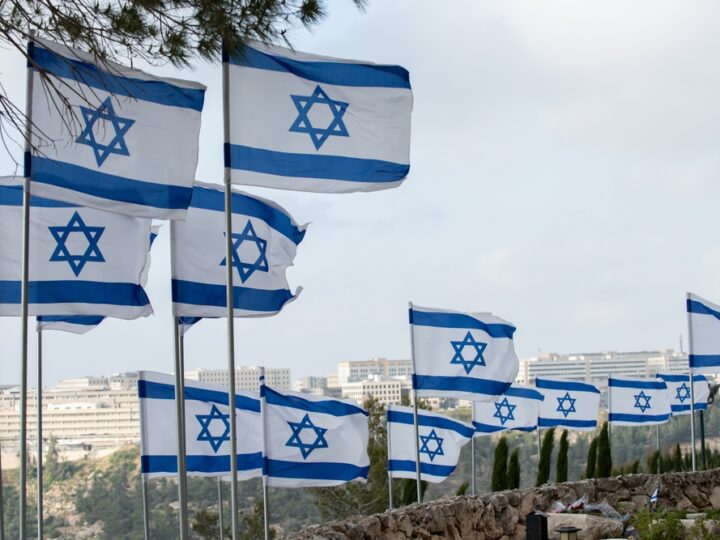 Multiple Israeli flags with blue Stars of David and horizontal blue stripes are flying on flagpoles against a backdrop of buildings and trees under a partly cloudy sky.