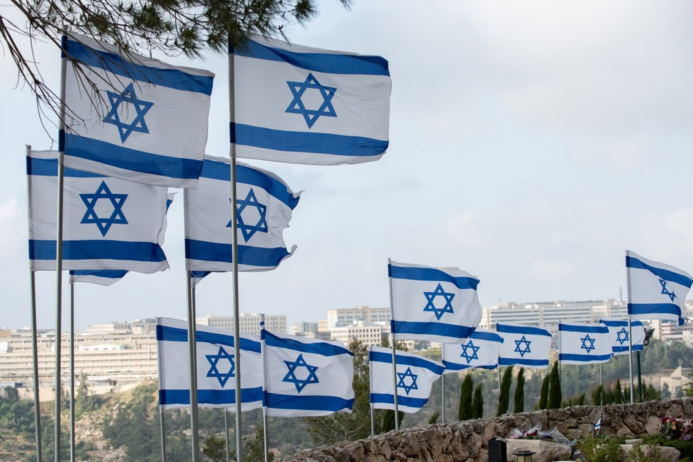 Multiple Israeli flags with blue Stars of David and horizontal blue stripes are flying on flagpoles against a backdrop of buildings and trees under a partly cloudy sky.