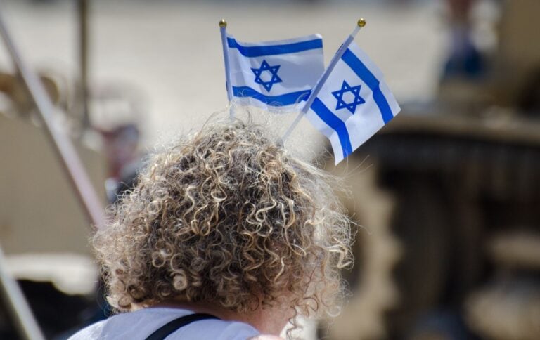 A woman with curly hair wears two small Israeli flags on their head, attached to a headband. The background appears outdoors and is out of focus.