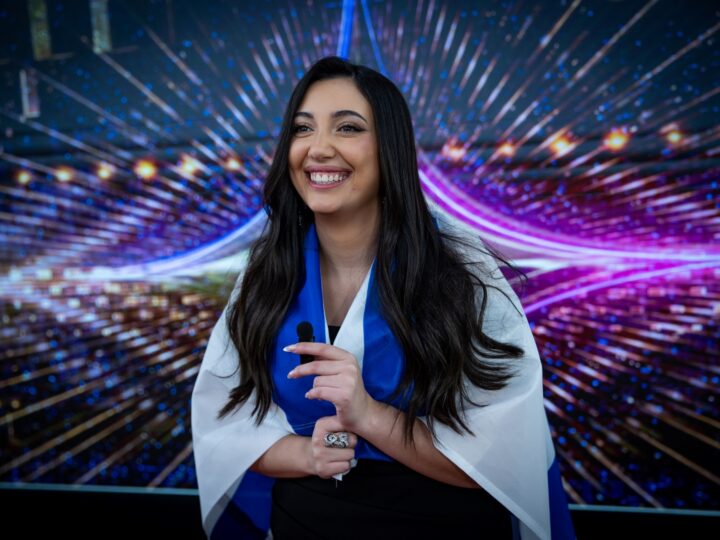 A photo of Yuval Raphael smiling and holding a microphone, draped in the white and blue Israeli flag . She stands in front of a vibrant, starburst-patterned backdrop with blue and purple lights.