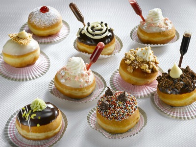 A spread of nine hanukkah donuts set on a white table, they each have different color frostings and different toppings representing a variety of flavors, including powdered sugar, chocolate frosting, jelly, pistachio cream, sprinkles and cookies. Each donut sits on a colorful cupcake wrapper laid flat on the table. 